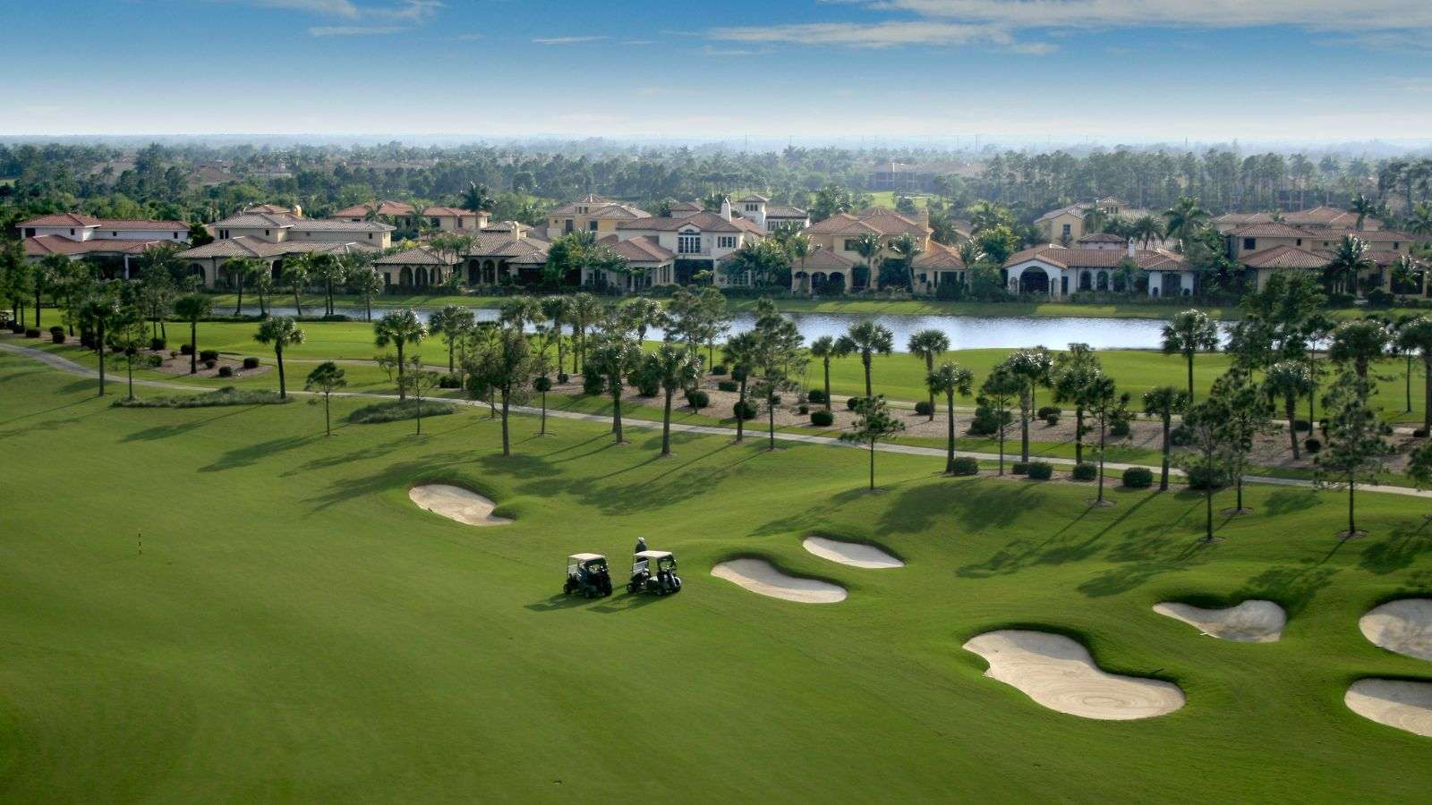 Aerial view of a Florida golf course community with two golf carts, green fairways, palm trees, a lake, and single-family homes in the distance