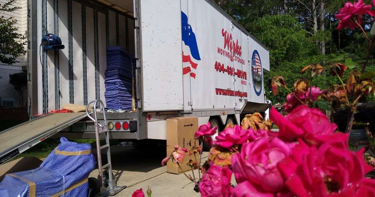 Wirks Moving & Storage moving truck in front of a customer’s home with the trailer open and the ramp down. Furniture wrapped in moving blankets, a packed box, and a moving dolly are placed nearby.