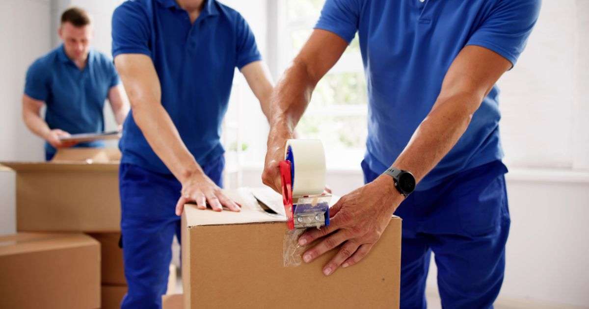 A team of three movers packing boxes in a customer’s home, sealing boxes, and preparing an inventory of household goods.