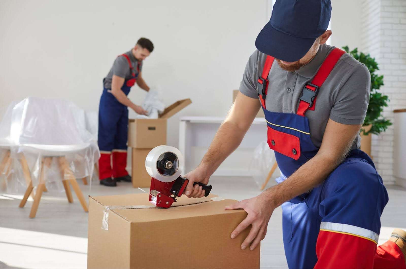 Flowery Branch mover in blue and red overalls taping up a box while another mover packs a box in the background. 