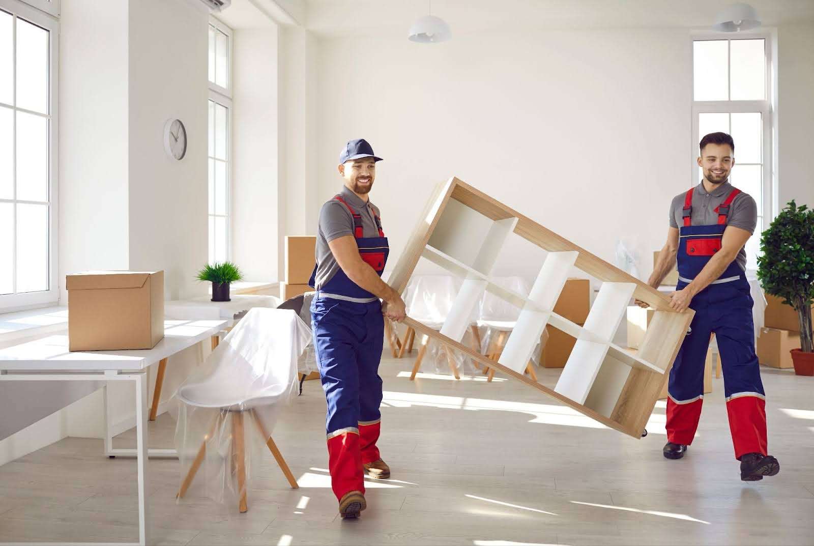 Two Flowery Branch movers in overalls smiling and carrying a shelf out of a home 