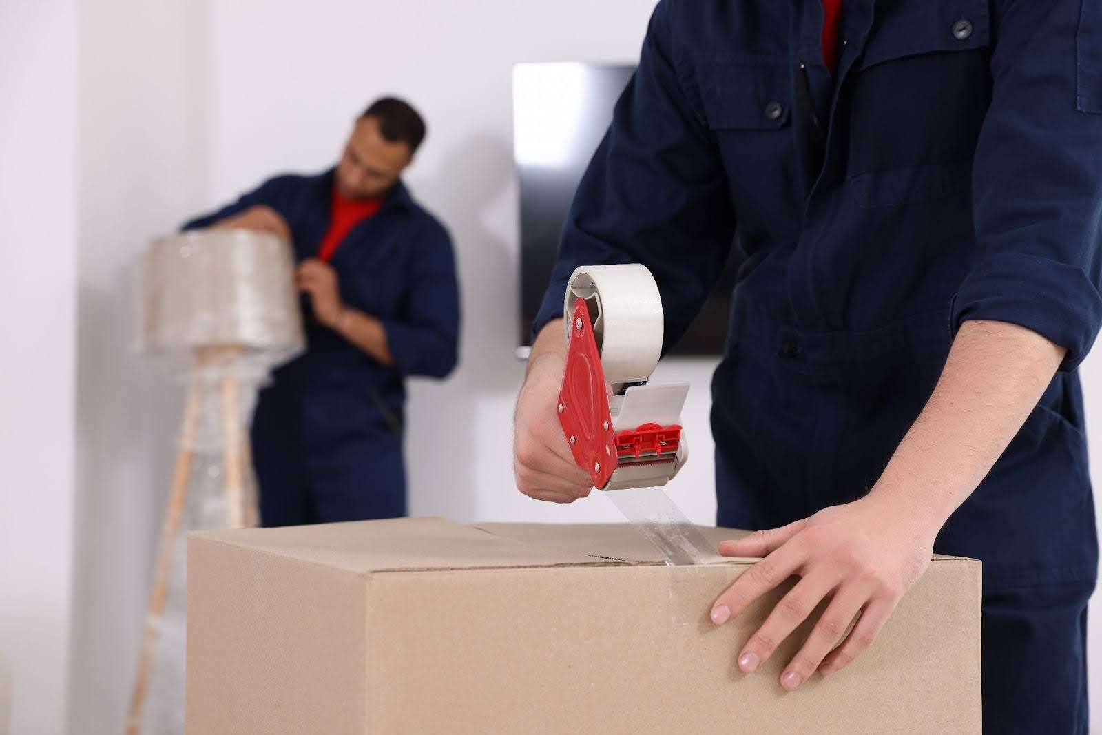 Close up shot of mover taping up a box while another mover carefully wraps a lamp in plastic wrap in the background.