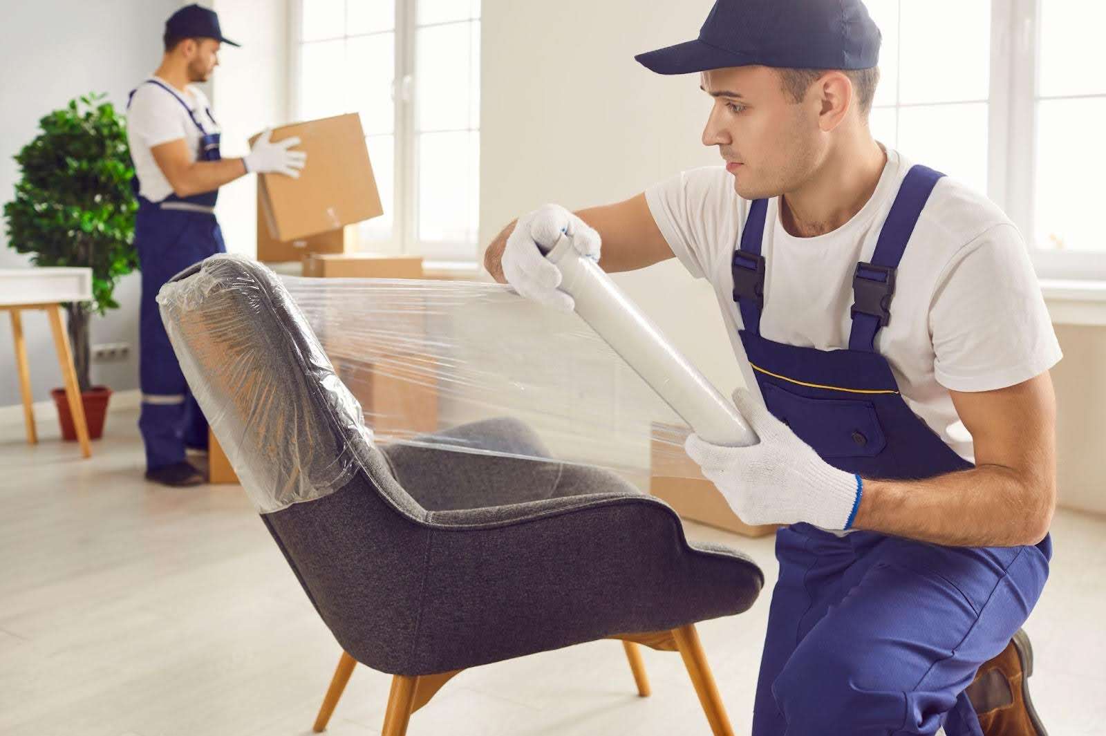 Mover with blue overalls and white gloves wraps a chair in plastic wrap while another mover moves boxes in the background. 