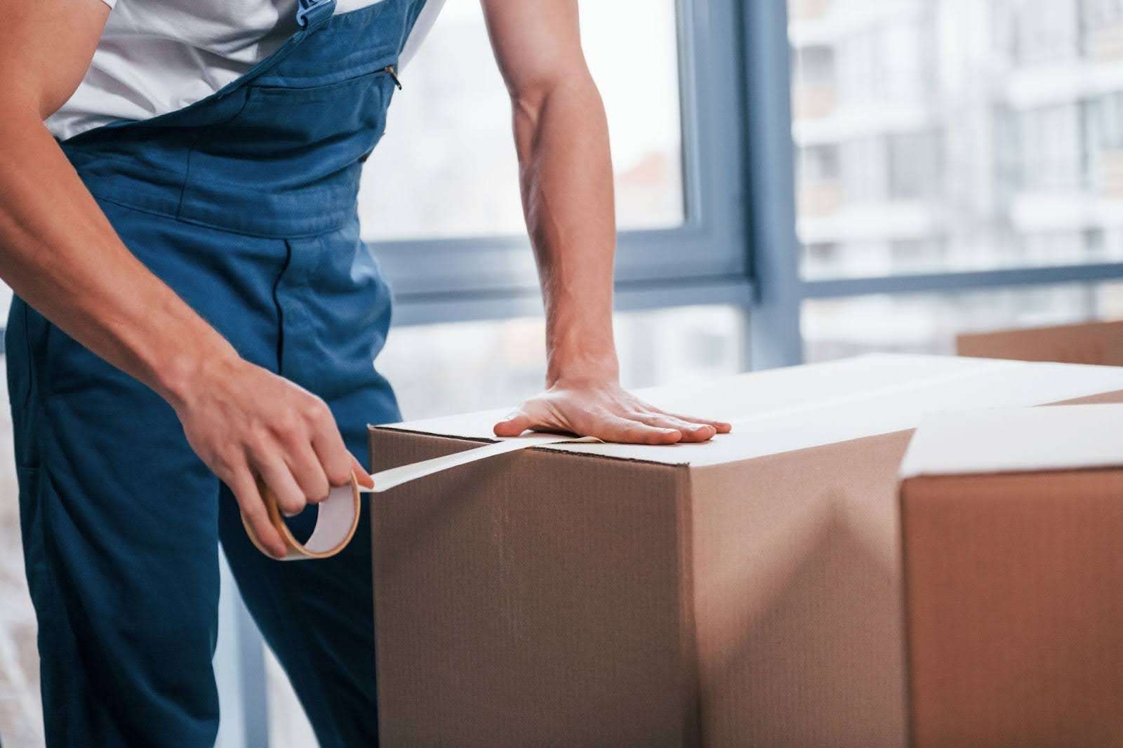 Mover in blue overalls putting tape on a cardboard box
